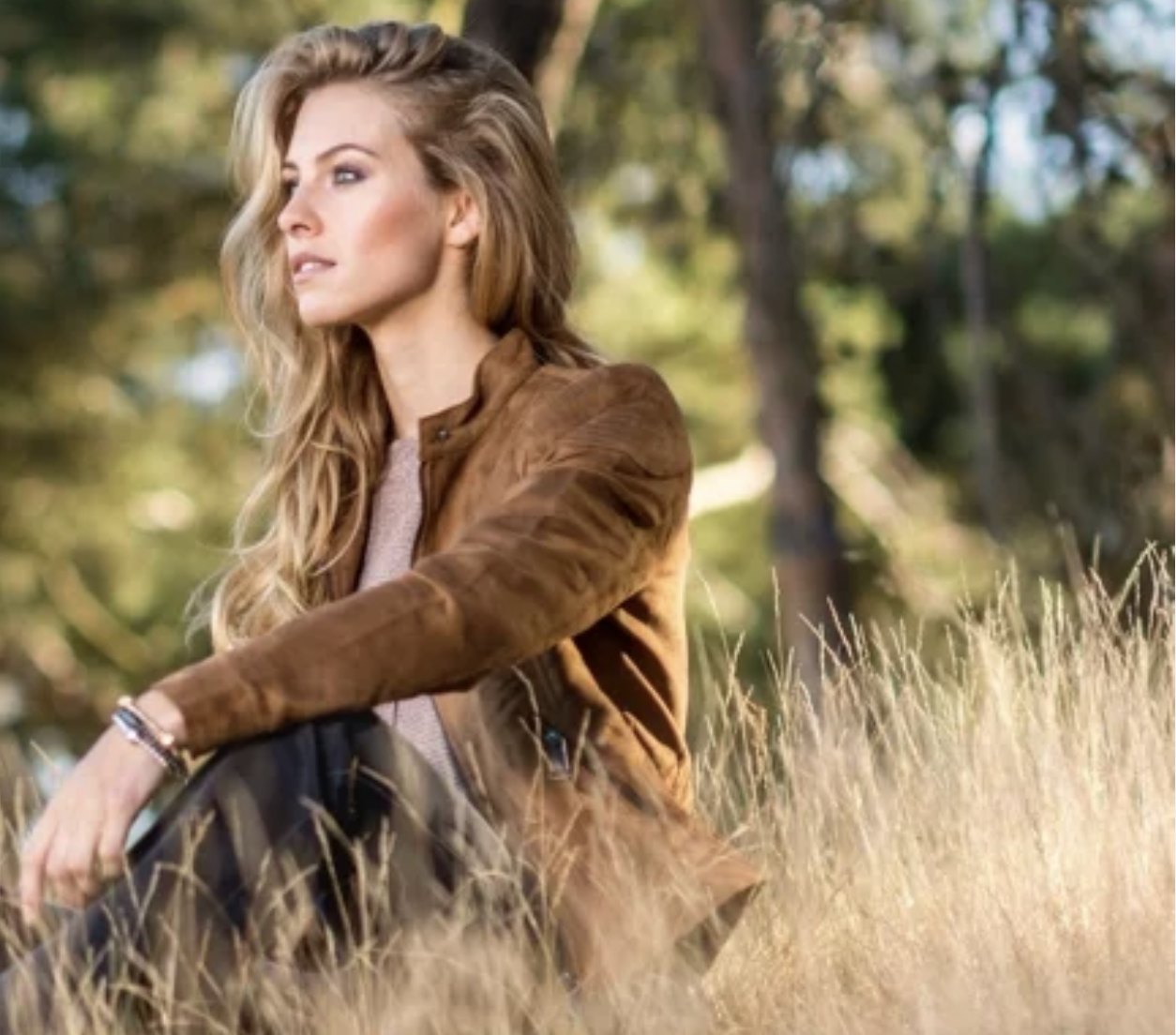 Model in suède jas zittend in hoog gras in een dennenbos, avondlicht